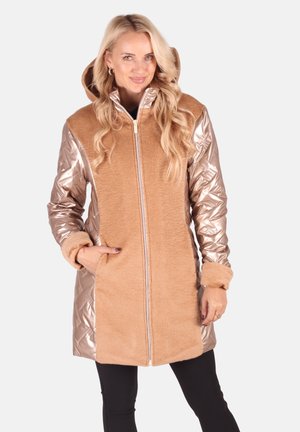 Frau in einem beigen Kapuzenmantel mit glänzenden, gesteppte metallischen Ärmeln, die mit einer Hand in der Tasche steht und lächelt.