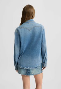 Camicia in denim blu chiaro con maniche lunghe, colletto a punta e orlo strappato. Presenta cuciture in evidenza e un logo discreto vicino al colletto.