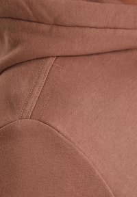 Un sweat en matériau beige clair avec une texture douce, doté d'une capuche et d'une couture contrastante le long des coutures.