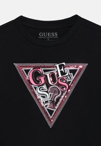 T-shirt en coton bio noir avec un design de logo triangulaire en sequins métalliques roses et argentés, comprenant le mot "GUESS" et des accents en forme de point d'interrogation.