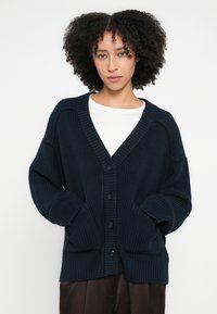 Tengerészkék kötött kardigán mély V-nyakkivágással, gombos záródással, bordázott mandzsettákkal és két elülső zsebbel. Texturált anyag és laza szabás.