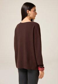 Suéter oversized color granate con escote redondeado, puños y dobladillo acanalados. Modelo lleva una pulsera en una muñeca. Material de punto suave.