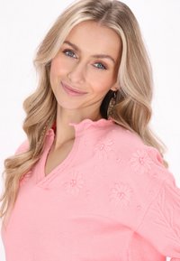 Maglione rosa lavorato a maglia con scollatura a V, presenta ricami floreali e un colletto arricciato. Texture morbida con dettagli in rilievo discreti.