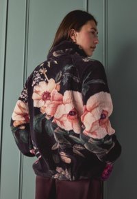 Sötét tónusú, virágmintás polárdzseki, puha textúrával, magas gallérral, és élénk rózsaszín és krémszínű virágokkal az ujjakon és a háton.