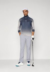 Golfoutfit mit einem marineblauen und grauen, verlaufenden Zip-up-Oberteil, hellgrauen Hosen und weißen Golfschuhen mit blauen Akzenten. Golfschläger in der linken Hand gehalten.