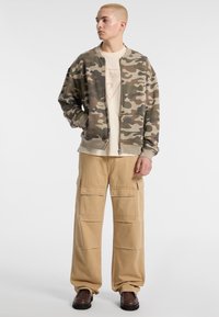 Camouflage bomberjack met rits, beige t-shirt met grafische print, losse beige cargobroek en bruine schoenen.