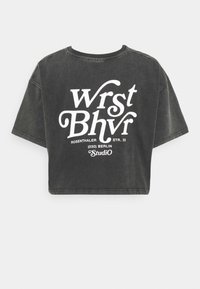Černé zkrácené tričko vyrobené ze 100% bavlny. Na přední straně se nachází velký bílý text se slovy "Wrst Bhvr" a detaily umístění v tučném, stylizovaném písmu.