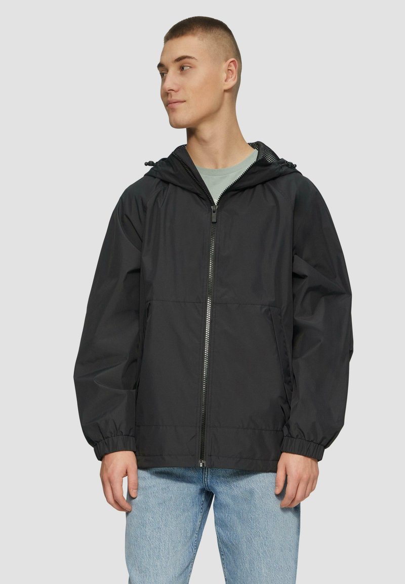 Zwarte hooded windbreakerjack met een ritsvoorkant, elastische manchetten en een relaxte pasvorm, gecombineerd met lichtblauwe jeans.