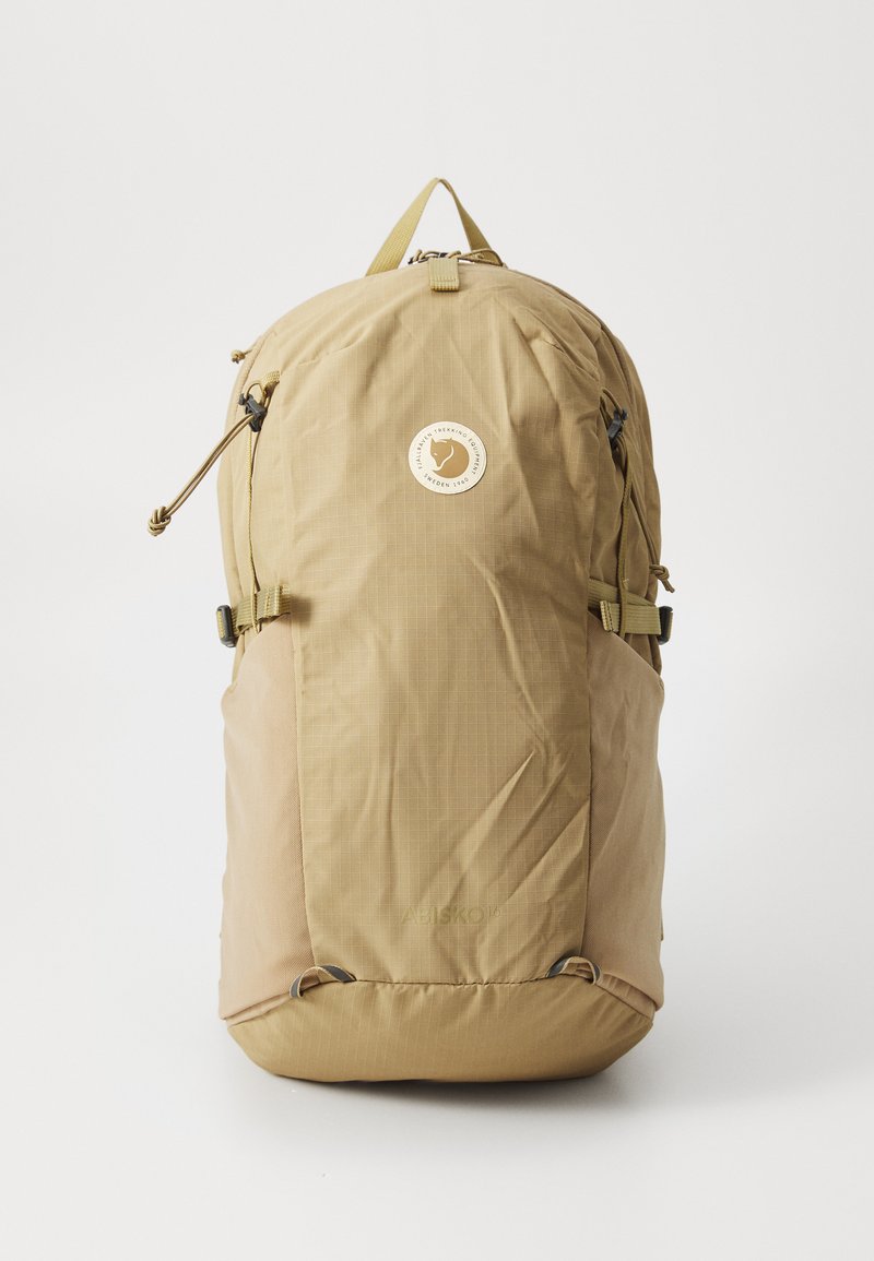 Beige backpack with a textured fabric, featuring a front logo patch, adjustable straps, and drawstring closure at the top.