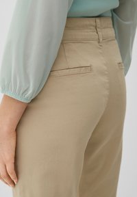 Beiga högmidjade byxor med en slät textur, med två bakfickor och en figursydd silhuett, tillsammans med en ljusblå blus.