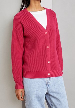 Fuchsia ribbet cardigan med V-hals, der har fem knapper, lange ærmer og en tætsiddende kant. Båret over en hvid T-shirt og lyse jeans.