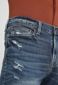 Džínové kalhoty v středně modrém odstínu s odřeninami, včetně roztržených látkových záplat a klasického pětipocket designu.