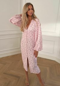 Femme aux longs cheveux blonds portant une robe midi rose à imprimé floral, avec des boutons sur le devant, des manches longues bouffantes, et des talons ouverts beiges.