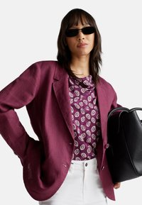 Blazer en lin bordeaux sur un haut violet à motifs, associé à un pantalon blanc et un sac fourre-tout noir texturé. Des lunettes de soleil complètent le look.