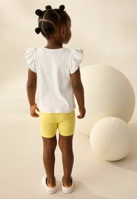 T-shirt bianca a costine con maniche a volant, abbinata a pantaloni corti gialli. Il bambino indossa scarpe bianche senza lacci, su uno sfondo neutro.