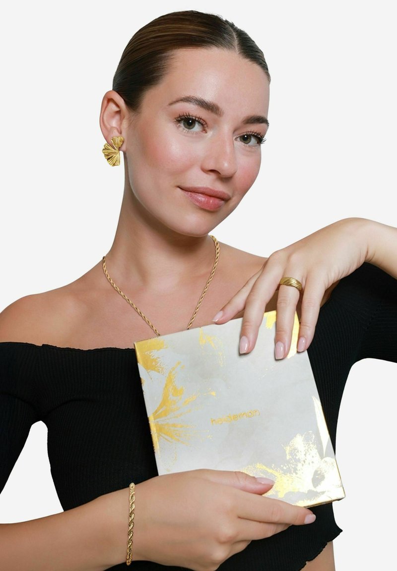 Goldschmuck mit einem strukturierten Design, darunter Ohrringe und ein Ring. Das Model hält ein eingepacktes Geschenk in einer gold-weißen gemusterten Schachtel.