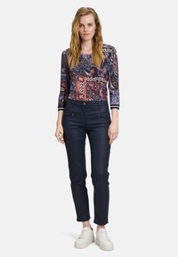 Langarmshirt mit einem mehrfarbigen Paisley- und geometrischen Muster; kombiniert mit dunkelblauen, eng anliegenden Hosen und weißen Sneakers.
