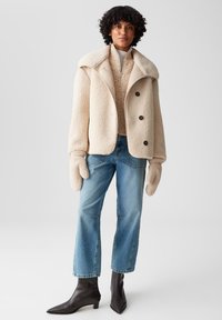 Beige Fleecejacke mit großem Kragen, schwarzen Knöpfen, kombiniert mit einem cremefarbenen Pullover, blauen geraden Jeans und spitzen schwarzen Stiefeletten.