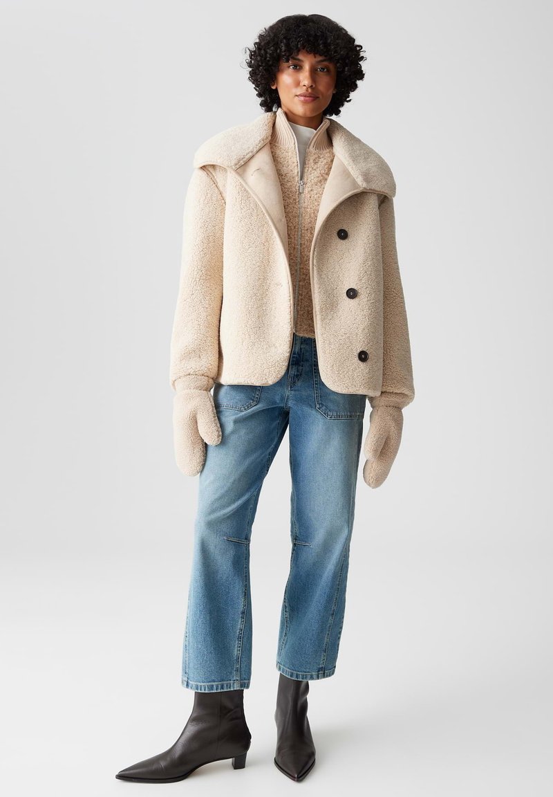 Beige Fleecejacke mit großem Kragen, schwarzen Knöpfen, kombiniert mit einem cremefarbenen Pullover, blauen geraden Jeans und spitzen schwarzen Stiefeletten.