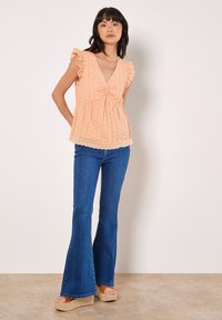 Blouse de couleur pêche avec un col en V, des manches à volants et un tissu texturé. Associée à un jean bleu évasé et des sandales à plateforme en espadrille.