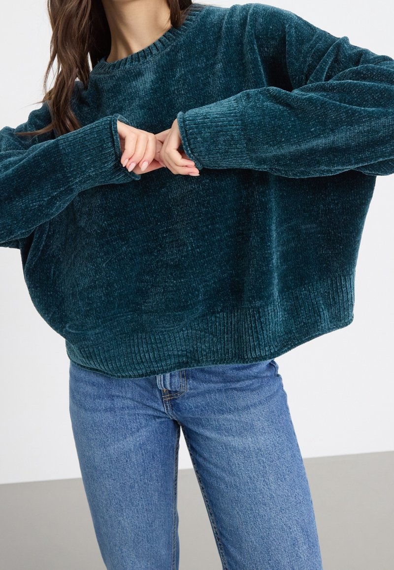 Femme portant un pull ample, vert foncé, côtelé et un jean bleu, ajustant les manches du pull avec ses mains.