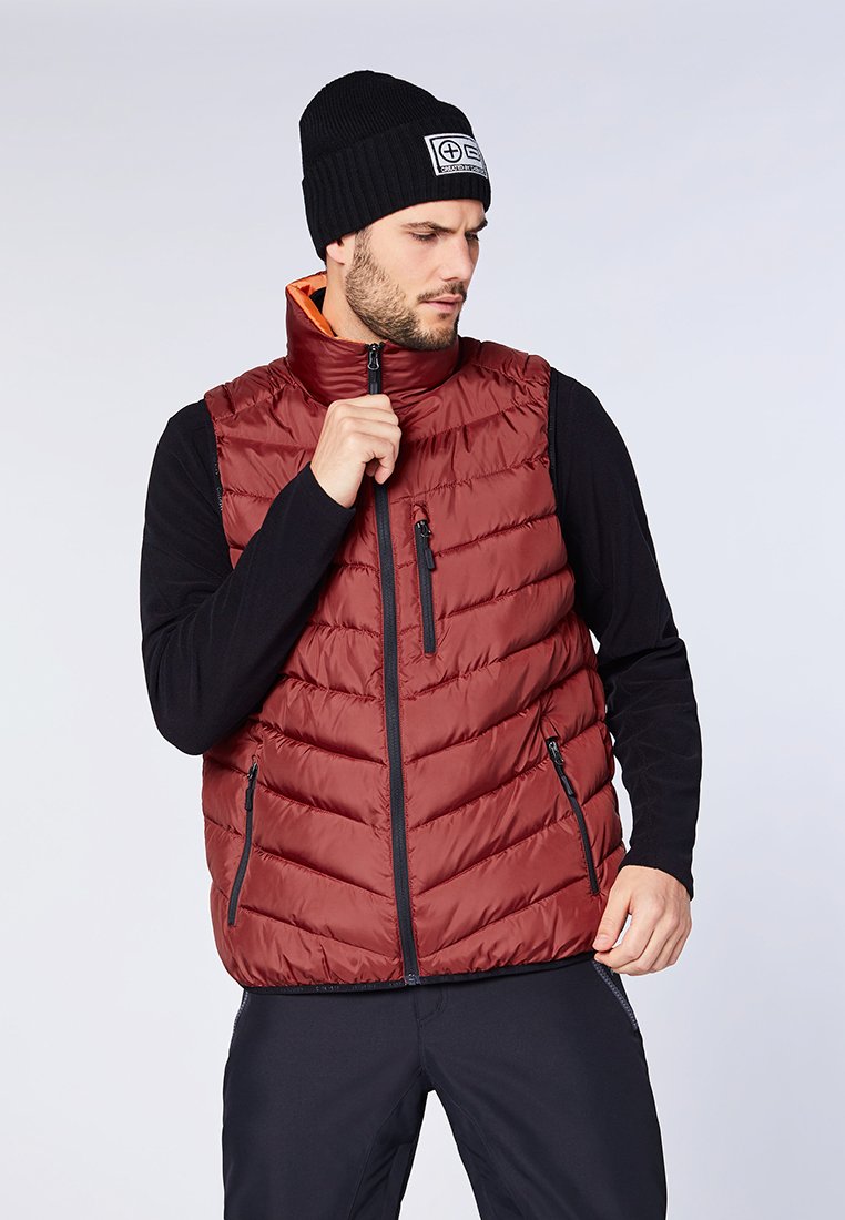 Burgunderfarbener gesteppter Weste mit hohem Kragen, Reißverschluss vorne und zwei Seitentaschen. Tragen über einem schwarzen Langarmshirt und kombiniert mit einer schwarzen Mütze.