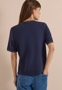Woman with brown hair wears navy blue short-sleeve top with animal print trim, blue jeans, standing with one hand in back pocket.