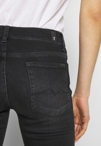 Personne portant un jean noir avec une poche arrière et un chemisier blanc rentré, montrant la taille et le bras droit détendu sur le côté.