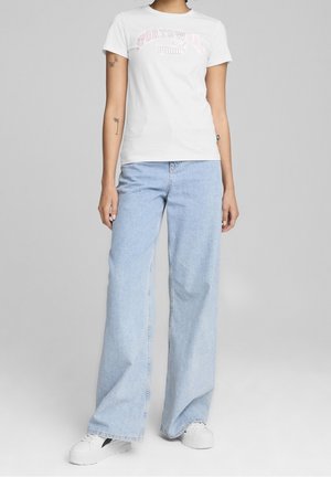 Bílé bavlněné tričko s růžovým nápisem "Sportswear Puma", doplněné světle modrými kalhotami širokých nohavic a bílými teniskami, stojící na šedém pozadí.