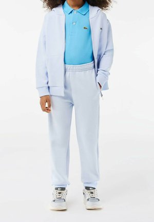 Enfant portant un sweat à capuche zippé bleu clair, un pantalon de survêtement assorti, un polo bleu et des baskets blanches, debout les mains détendues.