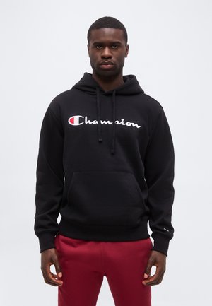 Mann trägt schwarzen Champion-Hoodie mit Fronttasche und roten Hosen, steht vor schlichtem hellem Hintergrund, schaut nach vorne.