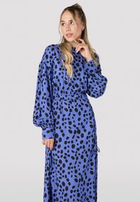 Robe à manches longues de couleur bleu vif avec un motif à taches noires. Présente une taille cintrée et un tissu fluide avec des poignets élastiques.