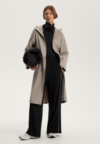 Hosszú bézs esőkabát kapucnival, fekete, cipzáras felső és hozzá illő, magas derekú nadrág felett viselve, egy fekete tárgyat tartva. Az outfittinget tornacipők egészítik ki.