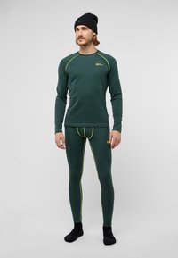 Haut à manches longues et bas thermiques verts avec des accents de couture jaunes. Design ajusté, texture côtelée, associés à des chaussettes noires et un bonnet.