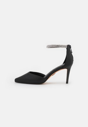 Sort spidse stilettohæle med et glitrende ankelstrop og lynlås bagpå mod en hvid baggrund.