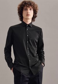Zwarte lange mouw blouse van gladde stof met een kraag, voorzien van een knoopsluiting aan de voorkant en een subtiele glans. Gecombineerd met een donkere broek.