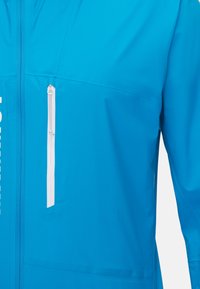 Veste légère bleu avec une poche poitrine zippée, texture lisse et design minimaliste avec des accents blancs.