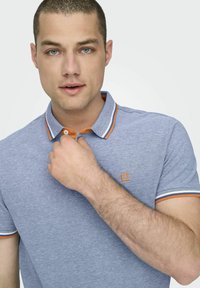 Polo de color azul hecho de tejido texturizado, con un collar a rayas en naranja y blanco. Logotipo en el lado izquierdo del pecho con un diseño cuadrado.