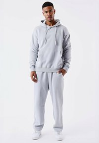 Felpa grigia chiara con cappuccio e pantaloni della tuta abbinati in tessuto morbido. La felpa presenta una tasca a marsupio e cordini. Indossata con sneakers bianche.