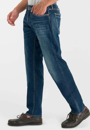 Man die donkerblauwe rechtvallende jeans en bruine leren bootschoenen draagt, lopend met één hand in de zak tegen een witte achtergrond.