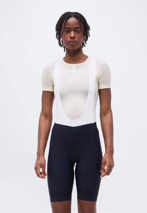 Persona de pie mirando al frente, vistiendo una camiseta beige de manga corta y pantalones cortos de ciclismo negros con tirantes blancos, sobre un fondo liso.