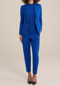 Kvinna iklädd en ljusblå kavaj, matchande skräddarsydda byxor, en stickad topp med textur och svarta spetsiga klackar som står mot en beige bakgrund.