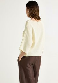 Lys gul strikket sweater med batwing ærmer, tekstureret stof og løs pasform, parret med brune bukser, vist fra ryggen.