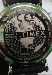 Stalowa tylna część zegarka Timex z grafiką globu i tekstem. Czarny materiałowy pasek z metalowymi akcentami. Wodoodporny do 50 m.