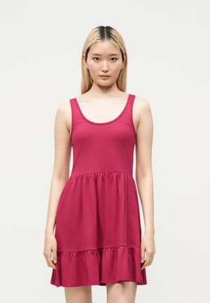 Jeune femme aux longs cheveux blonds portant une robe sans manches, longueur genoux, rose foncé, debout devant un fond blanc uni.