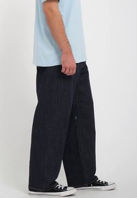 Jeans bleu foncé à jambes larges avec une texture en denim, ornés d'un accent de logo bleu, associés à un t-shirt bleu clair et des baskets noires.