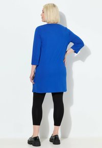 Blaue Tunika mit Dreiviertelärmeln und Seitenschlitzen, getragen über schwarzen Leggings. Das Model hat blonde Haare, die zu einem Bob geschnitten sind, und steht vor einem weißen Hintergrund.