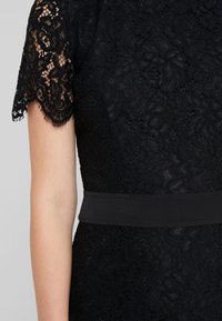 Schwarzes Spitzenkleid mit kurzen, gewellten Ärmeln, kunstvollen Blumenmustern und einem glatten schwarzen Taillenbund mit Nahtdetails.