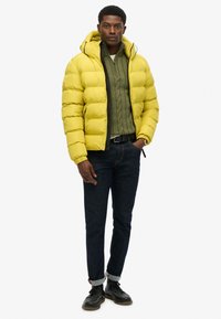 Geel pufferjack met een capuchon, voorzien van een rits aan de voorkant en zijzakken, gelaagd over een groene kabelgebreide trui en donkere denim jeans.