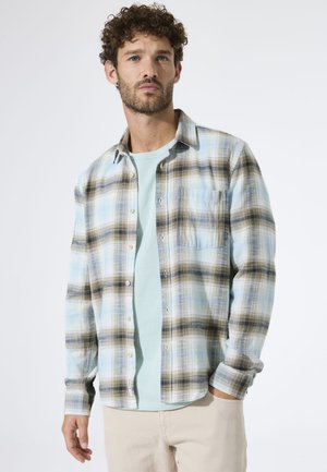 Kariertes Flanellhemd in hellblau, braun und weiß; Knopfleiste vorne und Brusttasche; getragen über einem hellgrünen T-Shirt.